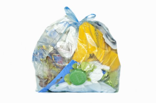 A transparent plastic rubbish bag filled with a variety of waste materials, including yellow and blue plastic packaging, a green plastic bottle cap, and crumpled paper or plastic items, securely tied at the top with a plastic tie. The bag appears to contain mixed household or commercial waste and is placed against a plain white background, illustrating typical waste collection for rubbish removal services. The contents indicate a mix of lightweight plastics and general refuse often handled by waste disposal specialists such as Commercial Waste Tadworth, serving areas around Tadworth and the KT postcode region. The image emphasizes the importance of proper waste containment for efficient rubbish removal and recycling efforts, relevant to the local waste management environment in northwest Surrey.