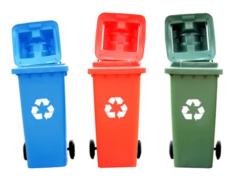 A row of three wheeled rubbish bins is displayed outdoors, positioned on a paved or concrete surface. The first bin on the left is blue with a hinged lid open at the top, revealing a textured interior for waste disposal, and features a white recycling symbol on its front. The middle bin is red with a similar hinged lid lifted, and also has a white recycling symbol visible. The third bin on the right is green, with its lid partially open, showing a textured interior and a white recycling symbol on the front. All bins have black wheels at the base, supporting their mobility, and sit against a neutral background, possibly a driveway or service area near a building in Tadworth or its surroundings. The lighting is natural, highlighting the vivid colors and clean surfaces of each bin, aligning with the waste management and rubbish removal services provided by Commercial Waste Tadworth, which serve the local area including postcode KT20.