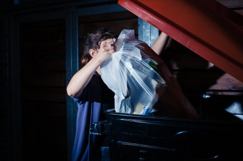 A young man with dark hair, wearing a dark short-sleeved shirt, is seen throwing a large, crumpled clear plastic bag filled with paper and cardboard waste into the top open compartment of a black skip or waste container. The skip is situated outdoors, possibly on a driveway or paved surface, with a dark wooden or composite fencing and blue-painted support beams visible in the background. The environment appears to be shaded or dimly lit, with the lighting focused on the waste disposal activity. This scene reflects a rubbish removal process typical of commercial waste services, such as those provided by Commercial Waste Tadworth, which operate in the Tadworth area of Surrey, postcode inference is KT20. The image captures the act of disposing of general waste materials, emphasizing the importance of proper rubbish handling and clearance in the context of local waste management services.