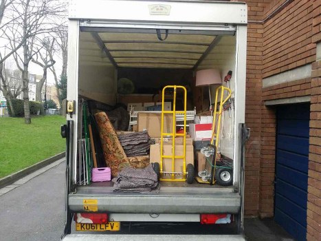 The image shows the open rear of a white commercial van parked on a residential street with a brick building to the right and a grassy area with leafless trees in the background. Inside the van, there is a mixture of household and miscellaneous rubbish, including a patterned armchair with fabric upholstery, a rolled-up carpet or large fabric item, and various smaller items stacked or leaning against the van's interior walls. A yellow hand truck is positioned upright near the back, and a small, purple plastic container is placed on the floor. The background reveals part of a suburban environment with paved pavement and some distant multi-storey buildings, indicating a residential or semi-urban area near Tadworth. The scene appears to be set during overcast weather, with natural light illuminating the contents clearly. This visual depicts typical waste collected by a rubbish removal service such as those offered by Commercial Waste Tadworth for local businesses or households.