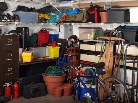 An indoor storage room filled with various household items and equipment, including plastic storage boxes, some with lids and others open, stacked on wire shelving units. The shelves hold tools, containers, and miscellaneous items such as a red bag, yellow buckets, and rolled-up cables. In the foreground, there is a blue bicycle with a black seat and a small black bag attached to the handlebars, positioned next to a wooden drying rack and several terra cotta plant pots of different sizes. To the left, a black filing cabinet and a black speaker are visible, along with a red fire extinguisher on the floor. The environment appears cluttered, with items typically associated with storage or garage organization, situated against a backdrop of peeling painted wall. The scene is lit by ambient light, emphasizing the variety of textures and materials including plastic, metal, and wood, all within a space that could be part of a property in the Tadworth area, supporting services by Commercial Waste Tadworth for rubbish removal and storage clearance.