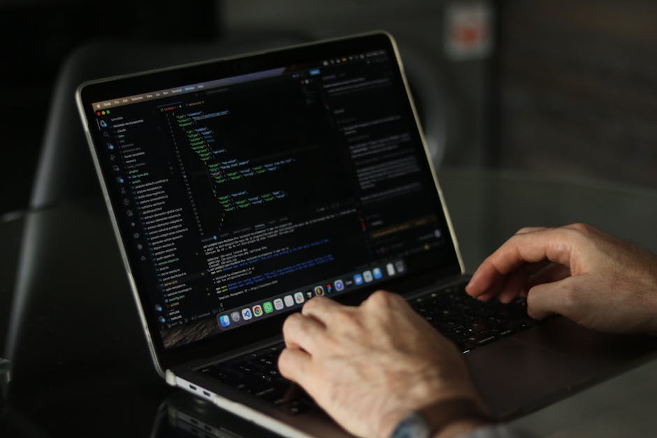 A pair of human hands with light skin tone and visible veins typing on a slender, silver laptop with a black keyboard, positioned on a dark grey desk or table. The laptop screen displays lines of code