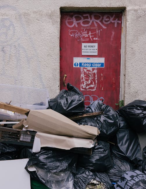 An outdoor scene showing a red metal fire exit door covered in graffiti, situated within a light beige, rough-textured wall. The door has multiple stickers, including a white one with the words 'No Mo